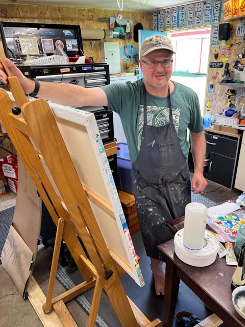 Eric Twait, standing in his art studio surrounded by empty and full canvases and supplies